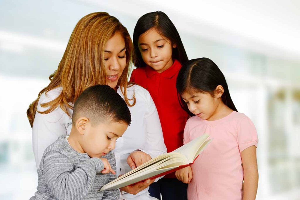 Hispanic woman reading a book to three hispanic children, 2 girls and 1 boy, ranging from pre-teen age to toddler.